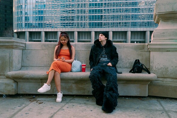 A woman in an orange dress and white shoes and a man in a black, furry outfit sit on a stone bench near the Chicago river. The woman is lighting a joint.