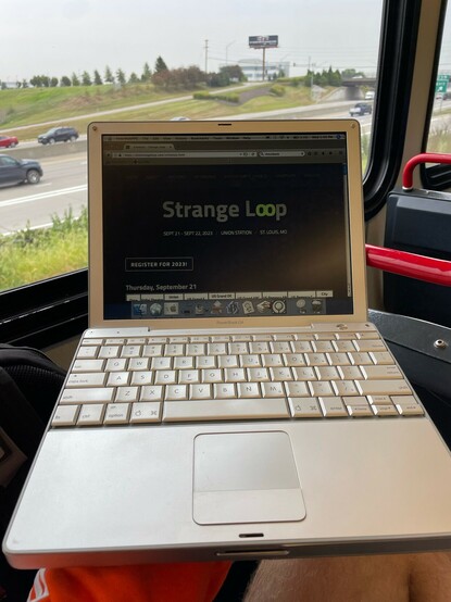 A 12" PowerBook G4 showing the Strange Loop home page in InterWebPPC, a modern web browser for PowerPC Macs. The window of a MetroLink train is visible behind it.