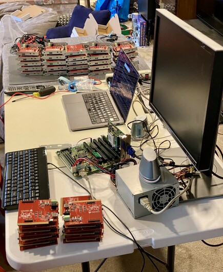 An overview of a folding table. In the foreground are stacks of plain PicoGUS boards, a 386 motherboard with PSU, keyboard, and monitor. In the middle is a MacBook used for firmware programming. In the background are stacks of PicoGUS PCBs with metal brackets attached. 