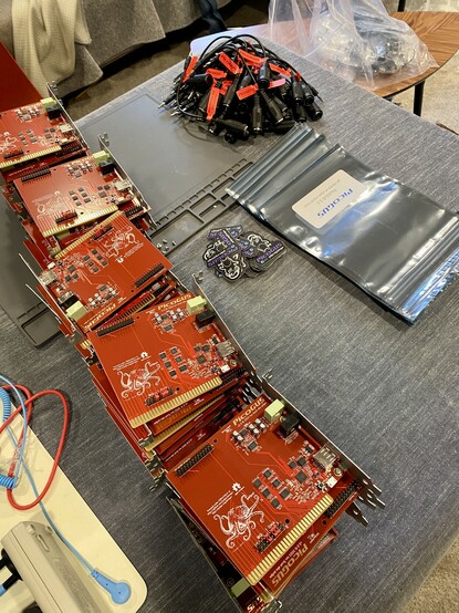 Stacks of assembled PicoGUS boards, along with a small pile of logo stickers, a stack of antistatic bags, and a pile of tagged MIDI adapter cables. 