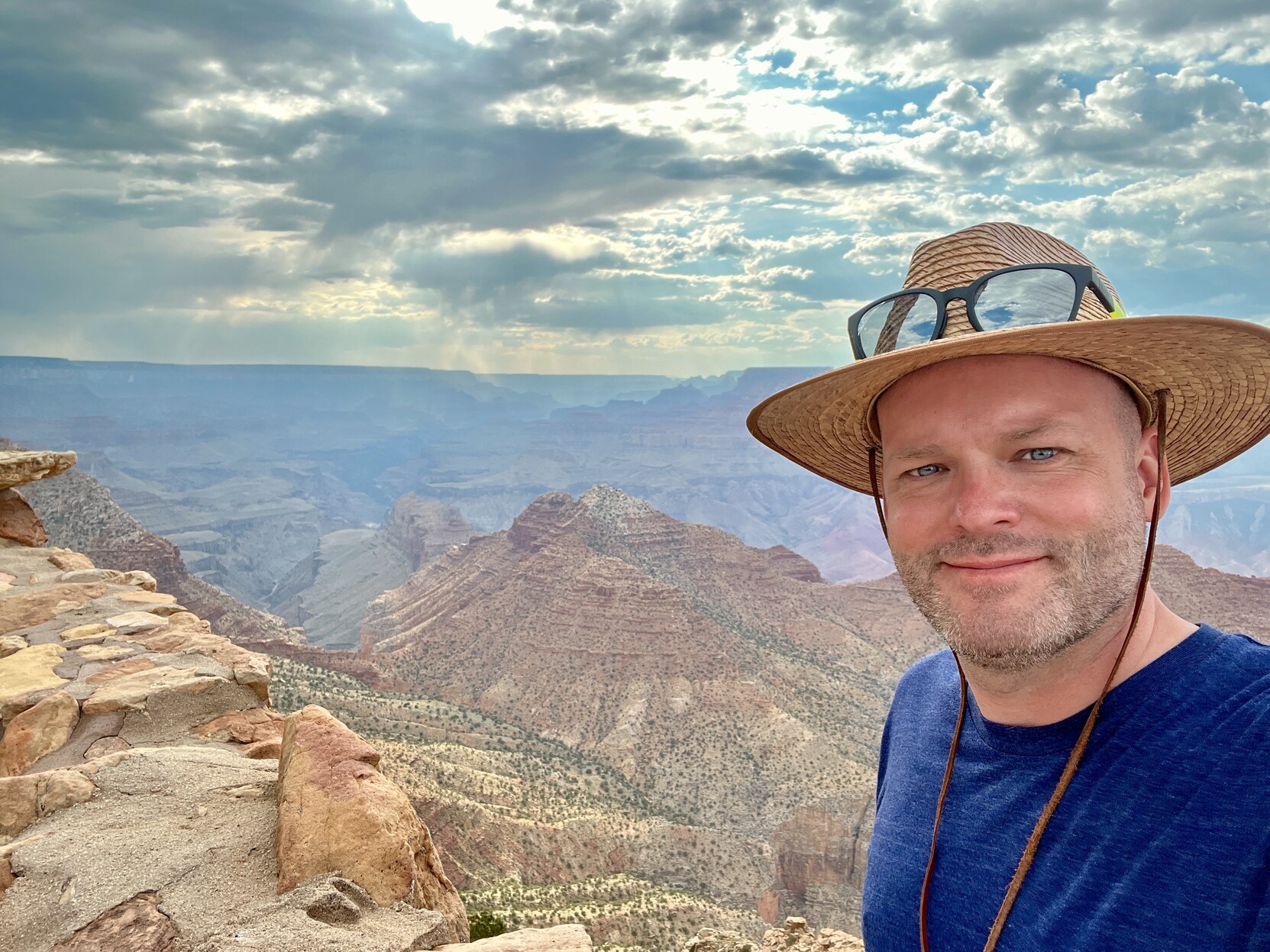 Selfie of me, a white middle aged guy with a slight smile. I’m wearing a straw hat with shades perched on the brim. The Grand Canyon is behind me, and you can see for miles. There’s rain out on the horizon. 