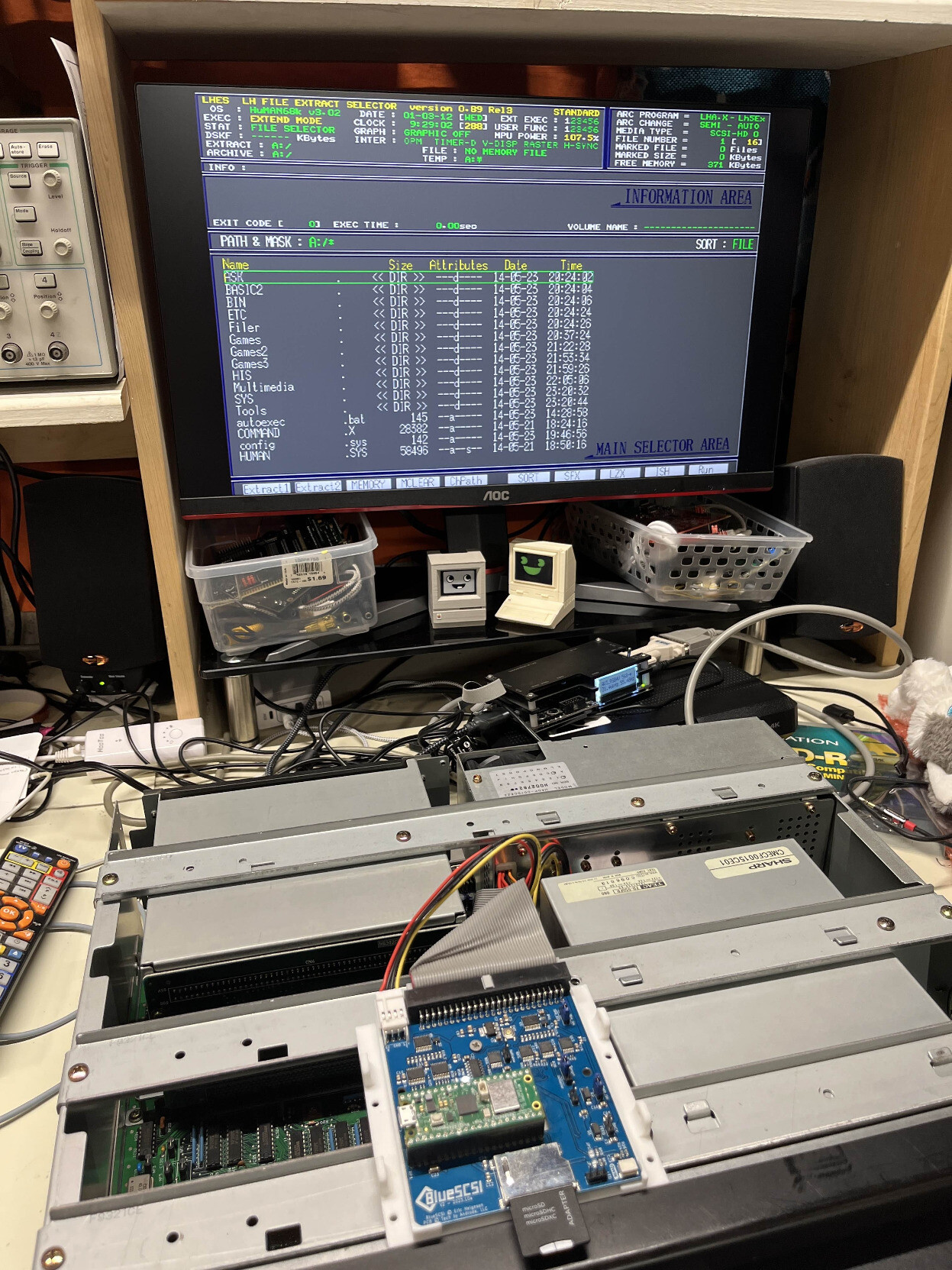 An open computer case, but not much of the internals are visible since it's blocked by large pieces of metal. There's a blue BlueSCSI PCB sitting on top connected to the computer with a ribbon cable. On the screen is a file selector program.