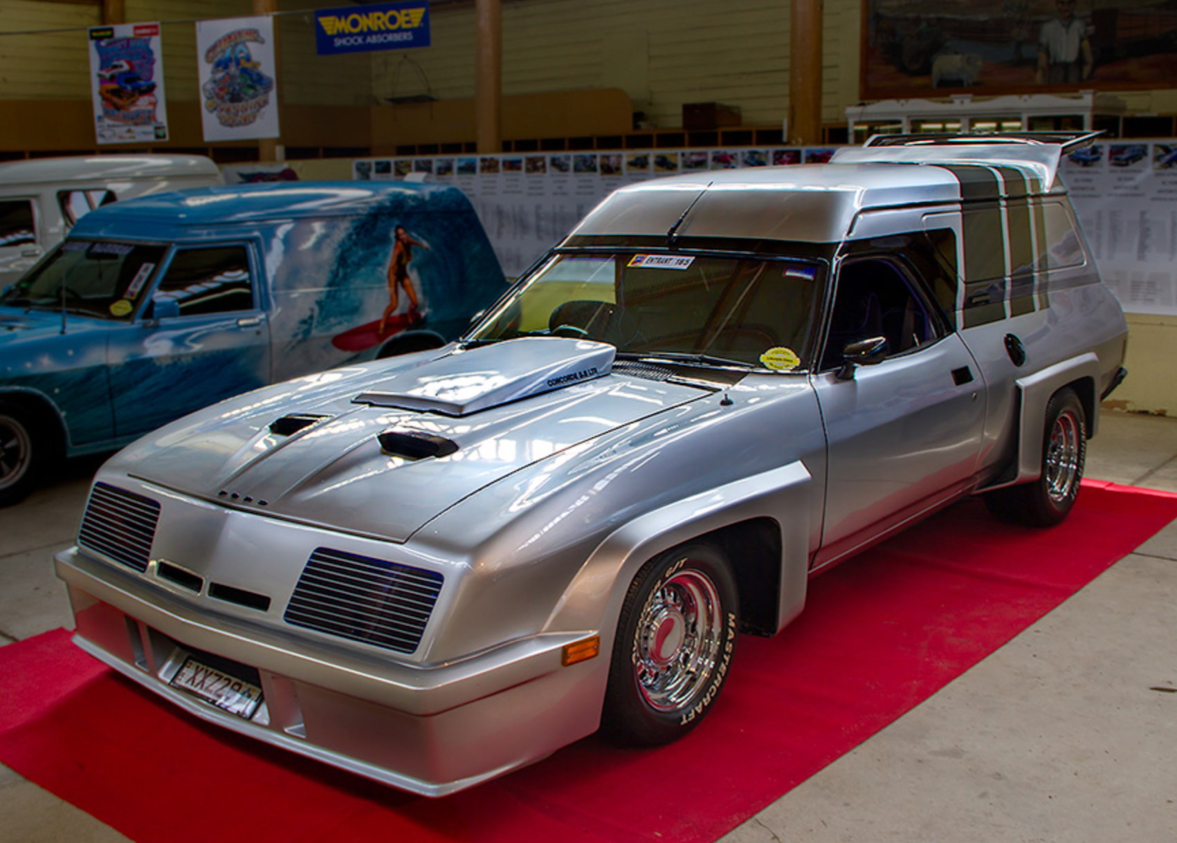 A photo of a silver Ford Falcon panelvan covered in bodykit parts and black stripes up over the back. It's seriously just slathered in fibreglass from the bonnet scoop, front bar, wheel arches, nostrils, rear spoiler, all over it just everywhere. It looks part 70s muscle car, part cheap sf movie prop.