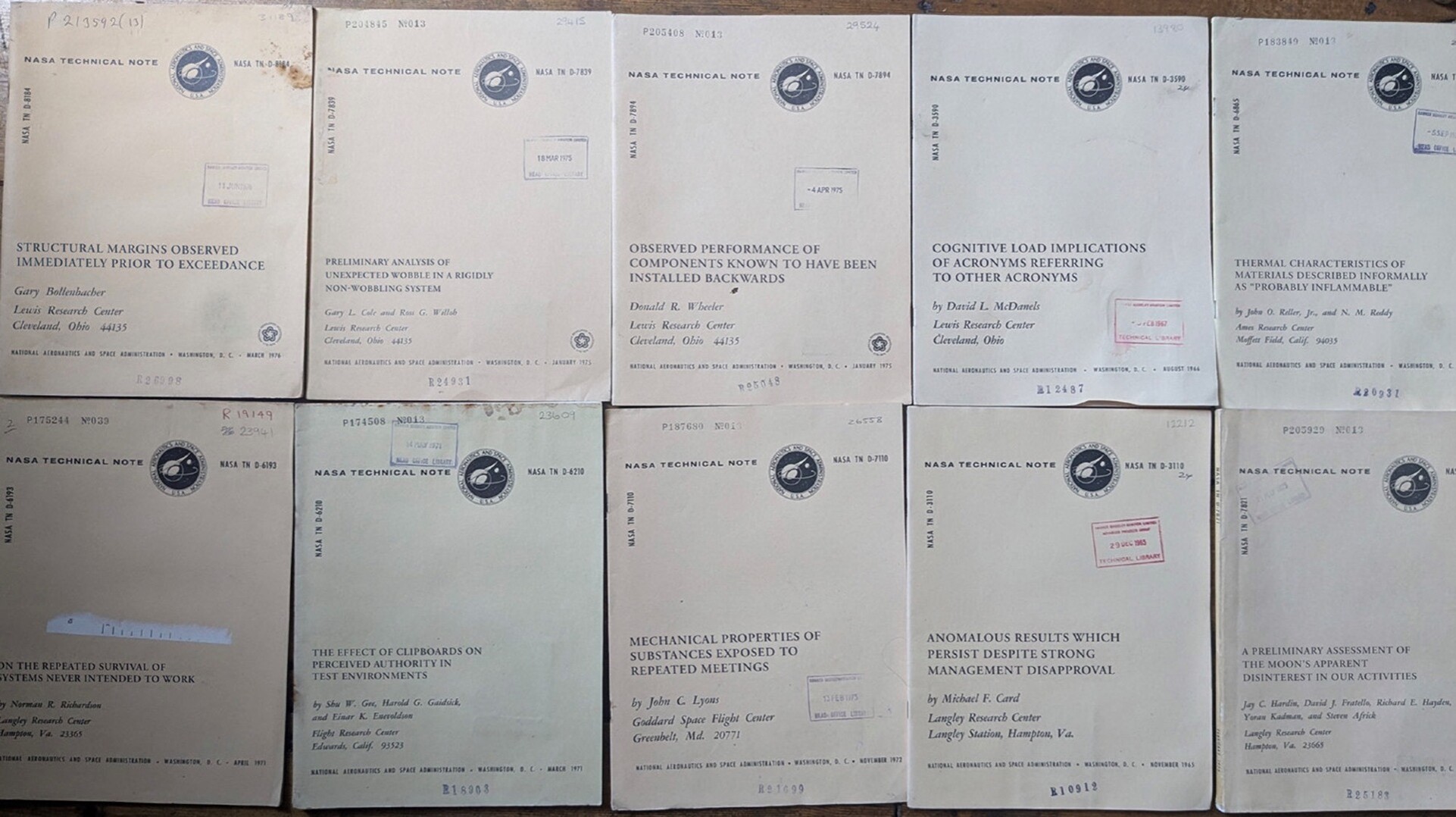 A photo of a bunch of NASA research documents, with titles like "STRUCTURAL MARGINS OBSERVED IMMEDIATELY PRIOR TO EXCEEDANCE", "PRELIMINARY ANALYSIS OF UNEXPECTED WOBBLE IN A RIGIDLY NON-WOBBLING SYSTEM", "OBSERVED PERFORMANCE OF COMPONENTS KNOWN TO HAVE BEEN INSTALLED BACKWARDS", "COGNITIVE LOAD IMPLICATIONS OF ACRONYMS REFERRING TO OTHER ACRONYMS", "THRMAL CHARACTERISTICS OF MATERIALS DESCRIBED AS 'PROBABLY INFLAMMABLE'", "ON THE REPEATED SURVIVAL OF SYSTEMS NEVER INTENDED TO WORK", "THE EFFECT OF CLIPBOARD ON PERCEIVED AUTHORITY IN TEST ENVIRONMENTS", "MECHANICAL PROPERTIES OF SUBSTANCES EXPOSED TO REPEATED MEETINGS", "ANOMALOUS RESULTS WHICH PERSIST DESPITE STRONG MANAGEMENT DISAPPROVAL", and "A PRELIMINARY ASSESSMENT OF THE MOON'S APPARENT DISINTEREST IN OUR ACTIVITIES"
